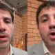 Homem com cabelo escuro e barba, vestindo terno claro, fala em frente a uma parede de tijolos