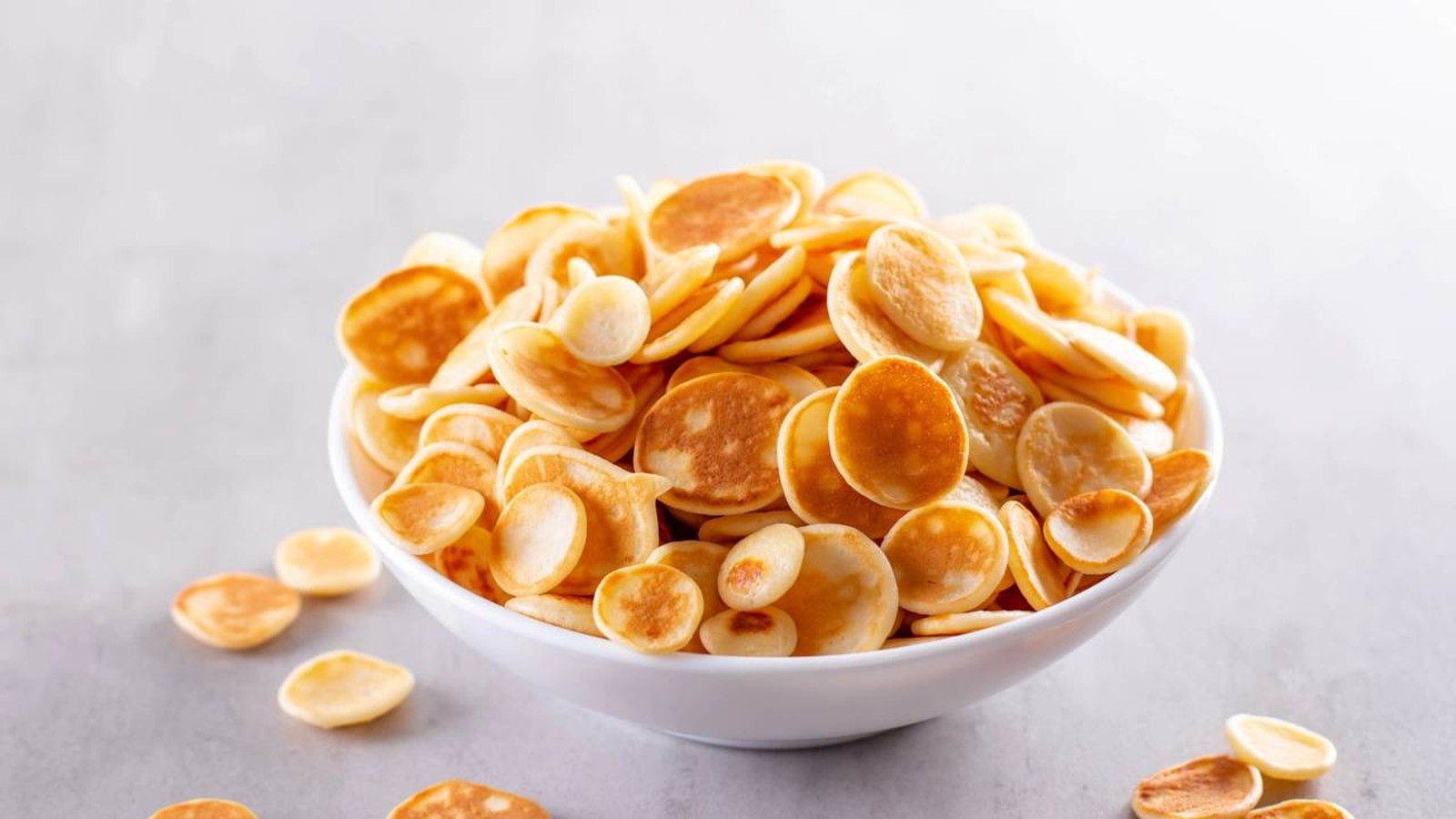 Tigela branca cheia de mini panquecas douradas e redondas sobre uma superfície cinza claro