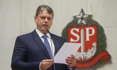 Tarcísio de Freitas de terno, discursa com microfone e papéis, brasão SP ao fundo