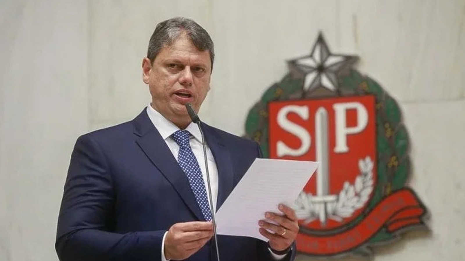 Tarcísio de Freitas de terno, discursa com microfone e papéis, brasão SP ao fundo