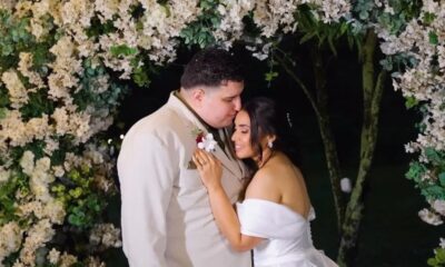 Homem de terno beija testa de mulher de vestido de noiva, sob arco floral
