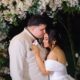 Homem de terno beija testa de mulher de vestido de noiva, sob arco floral