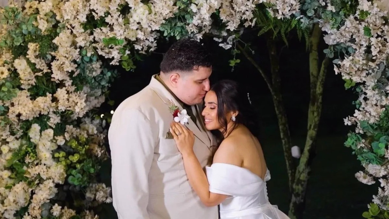 Homem de terno beija testa de mulher de vestido de noiva, sob arco floral