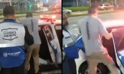 Homem com camiseta "CENOGRAFIA" sendo colocado em carro de polícia por agente "SEGURANÇA PRESENTE" à noite