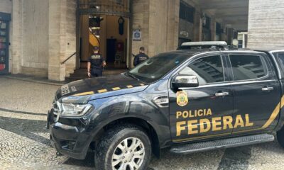 Viatura preta da Polícia Federal e dois policiais federais uniformizados em frente a um prédio