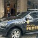 Viatura preta da Polícia Federal e dois policiais federais uniformizados em frente a um prédio