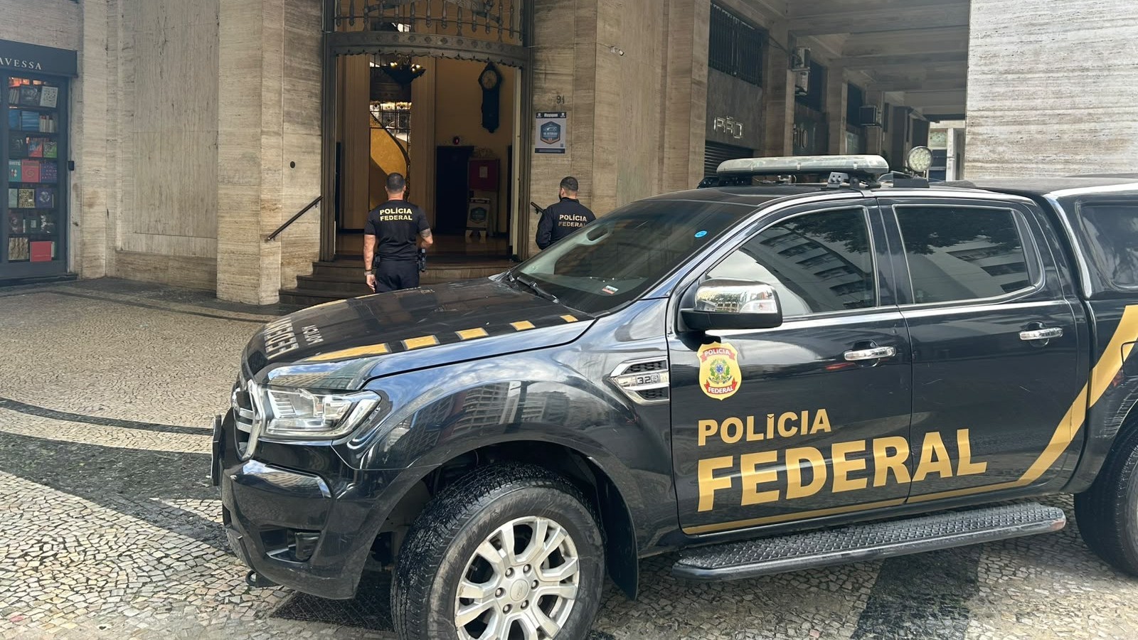 Viatura preta da Polícia Federal e dois policiais federais uniformizados em frente a um prédio