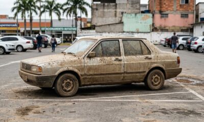 Carro sedan cinza coberto de lama estacionado, com outros carros, pessoas e prédios ao fundo.