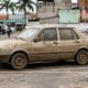 Carro sedan cinza coberto de lama estacionado, com outros carros, pessoas e prédios ao fundo.