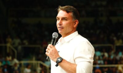 Flávio Bolsonaro de camisa branca e relógio, falando ao microfone para a plateia