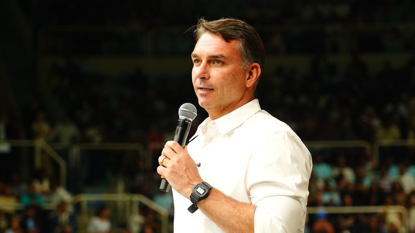 Flávio Bolsonaro de camisa branca e relógio, falando ao microfone para a plateia