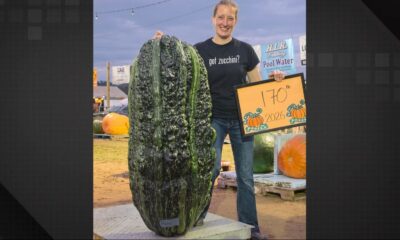 Mulher sorridente segura placa "170 lb 2026" ao lado de abobrinha gigante verde escura