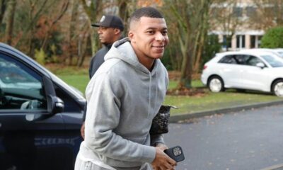 Kylian Mbappé sorrindo, usando moletom cinza, segurando smartphone, ao lado de carro escuro