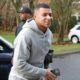 Kylian Mbappé sorrindo, usando moletom cinza, segurando smartphone, ao lado de carro escuro