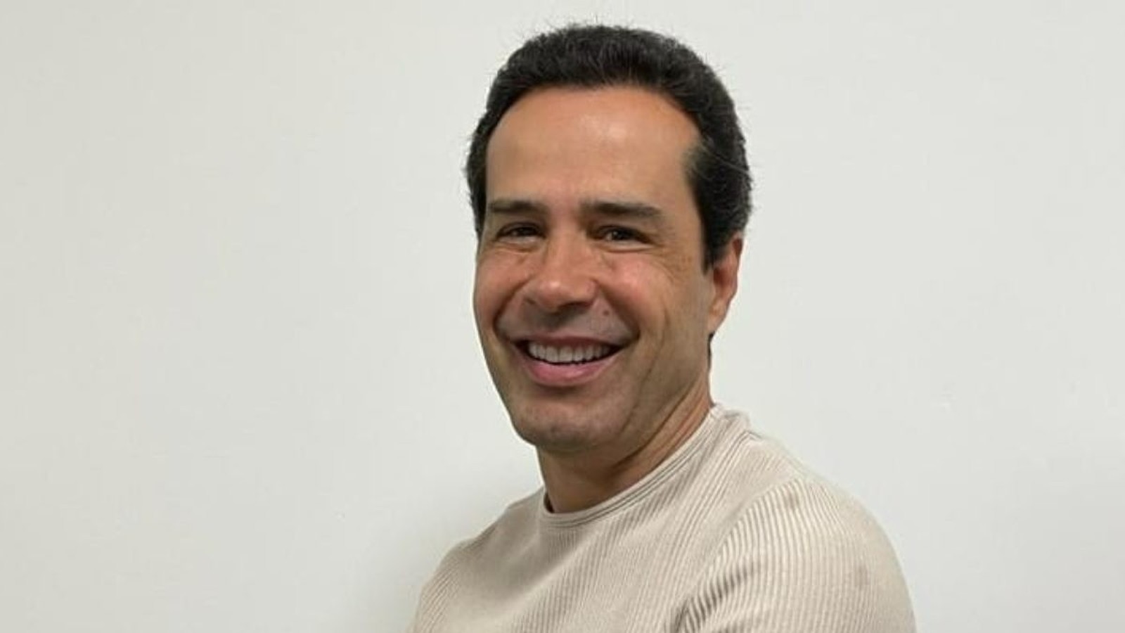 Homem sorridente de cabelo escuro, vestindo camisa clara, olhando para frente