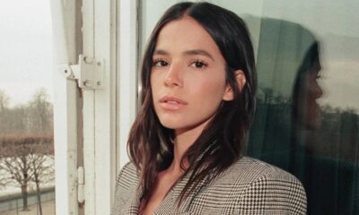 Bruna Marquezine, cabelo escuro, blazer xadrez, olhando à frente, com reflexo na janela