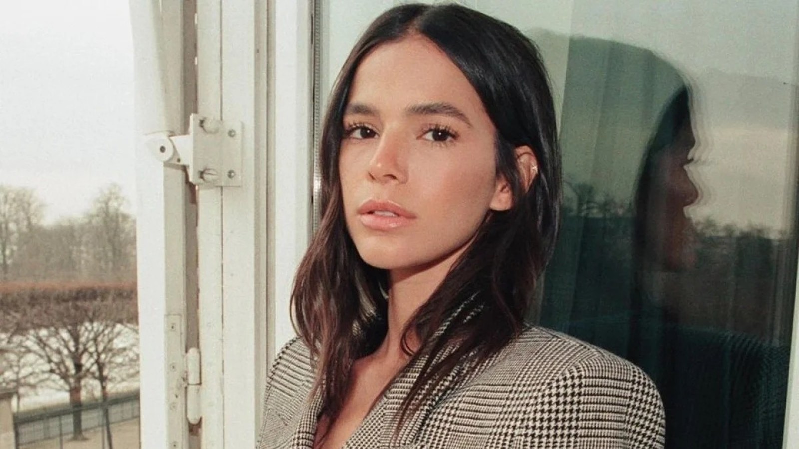 Bruna Marquezine, cabelo escuro, blazer xadrez, olhando à frente, com reflexo na janela