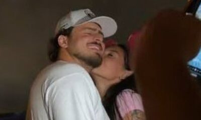 Jon Vlogs de boné branco sorrindo enquanto Ana Castela o beija na bochecha