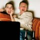 Casal aterrorizado no sofá assistindo TV, mulher e homem com bocas e olhos arregalados