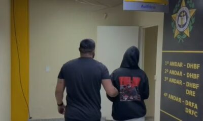 Agente escolta pessoa encapuzada em corredor de delegacia com placa de andares