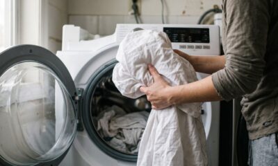 Pessoa com camisa cinza coloca lençóis brancos em máquina de lavar roupas com porta aberta