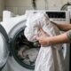 Pessoa com camisa cinza coloca lençóis brancos em máquina de lavar roupas com porta aberta