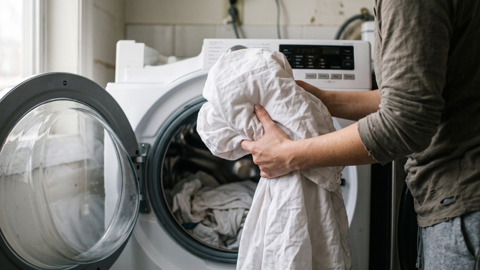 Pessoa com camisa cinza coloca lençóis brancos em máquina de lavar roupas com porta aberta