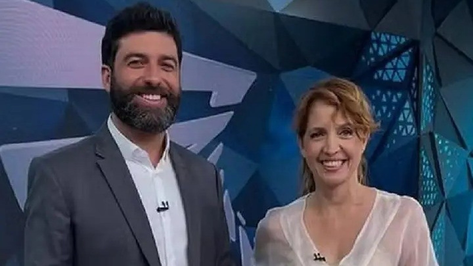 Rodrigo Carvalho e Poliana Abritta sorriem em estúdio de TV com fundo azul