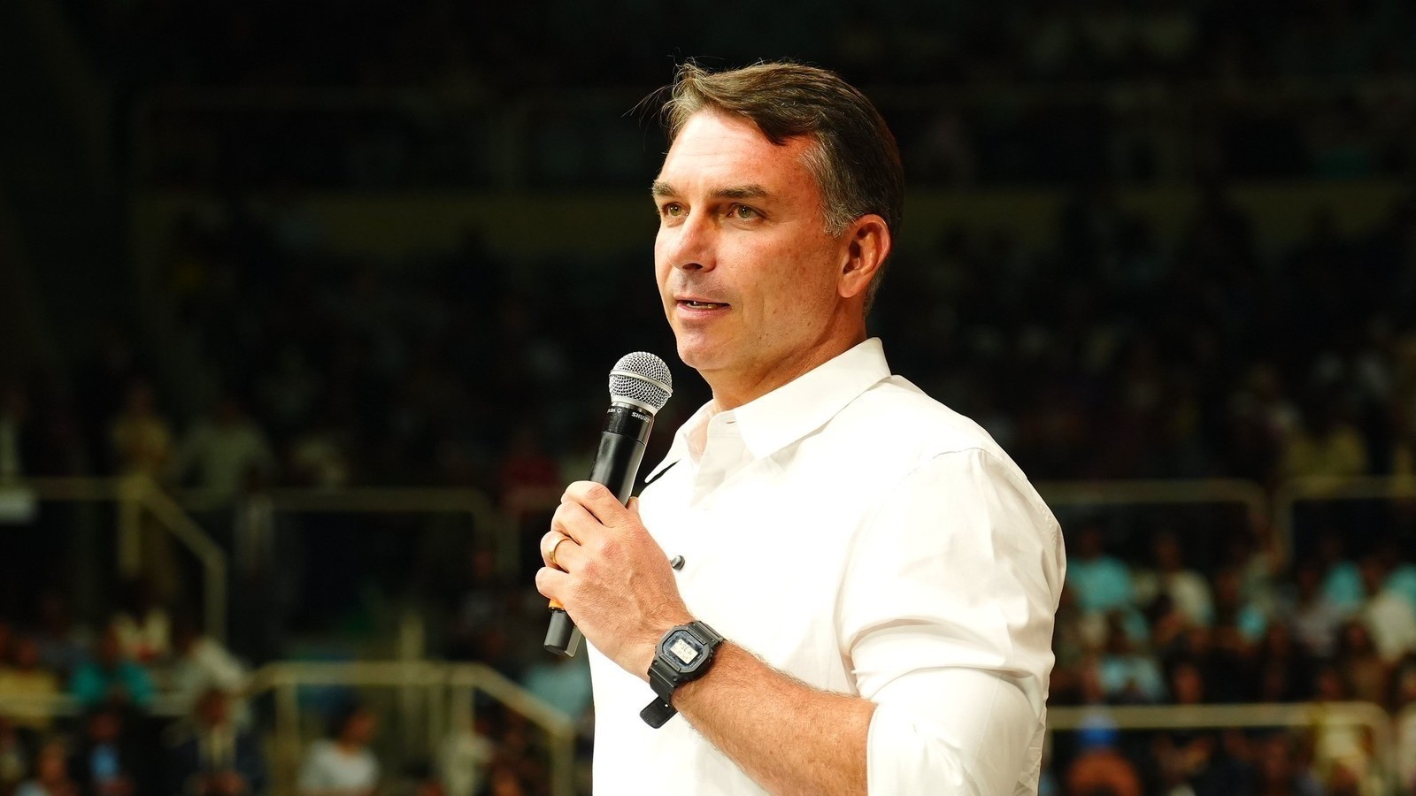 Homem de camisa branca, relógio digital preto, segurando microfone Shure, fundo de público desfocado