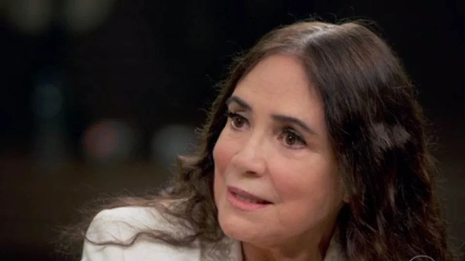 Mulher de meia-idade com cabelos escuros e grisalhos, usando blazer claro, olhando pensivemente