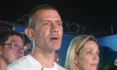 Homem de camiseta branca falando, com mulher loira ao lado e outro homem atrás, à noite