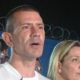 Homem de camiseta branca falando, com mulher loira ao lado e outro homem atrás, à noite