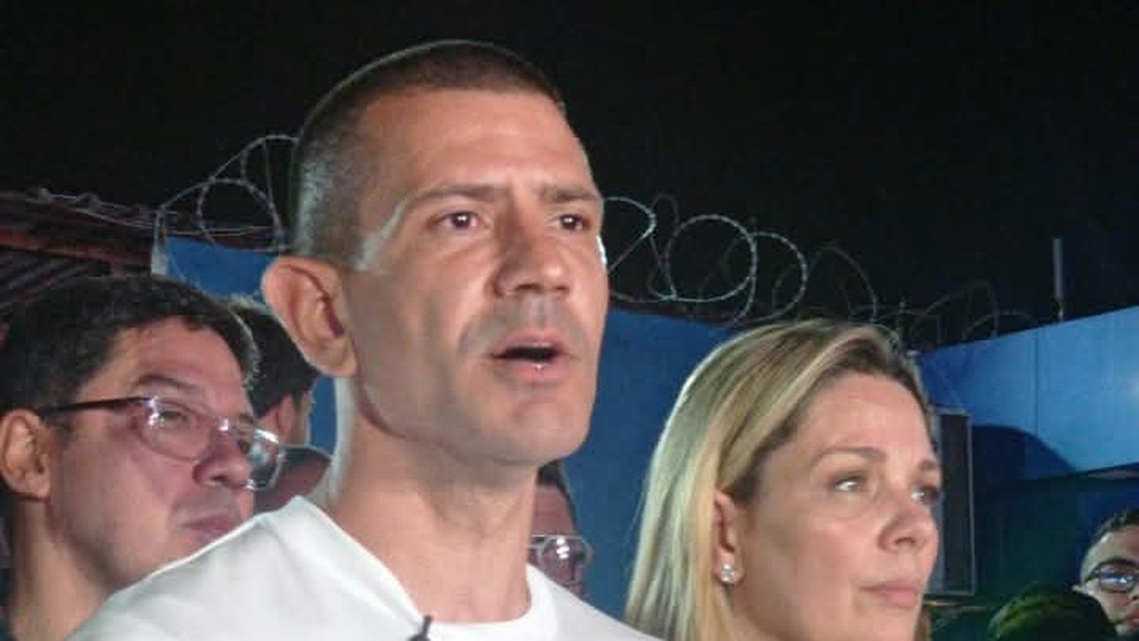 Homem de camiseta branca falando, com mulher loira ao lado e outro homem atrás, à noite