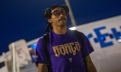 Homem com óculos amarelados, fones de ouvido e camisa roxa próximo a um avião