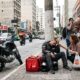 Entregador iFood exausto sentado na calçada, mulher agachada oferece água em rua movimentada