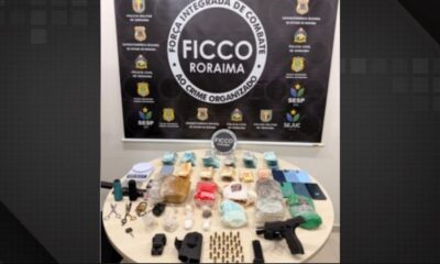 Mesa com drogas, dinheiro, pistola, munições e celulares apreendidos, com banner da FICCO Roraima