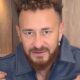 Homem com cabelo encaracolado e barba, vestindo jaqueta azul e branca, olhando para frente