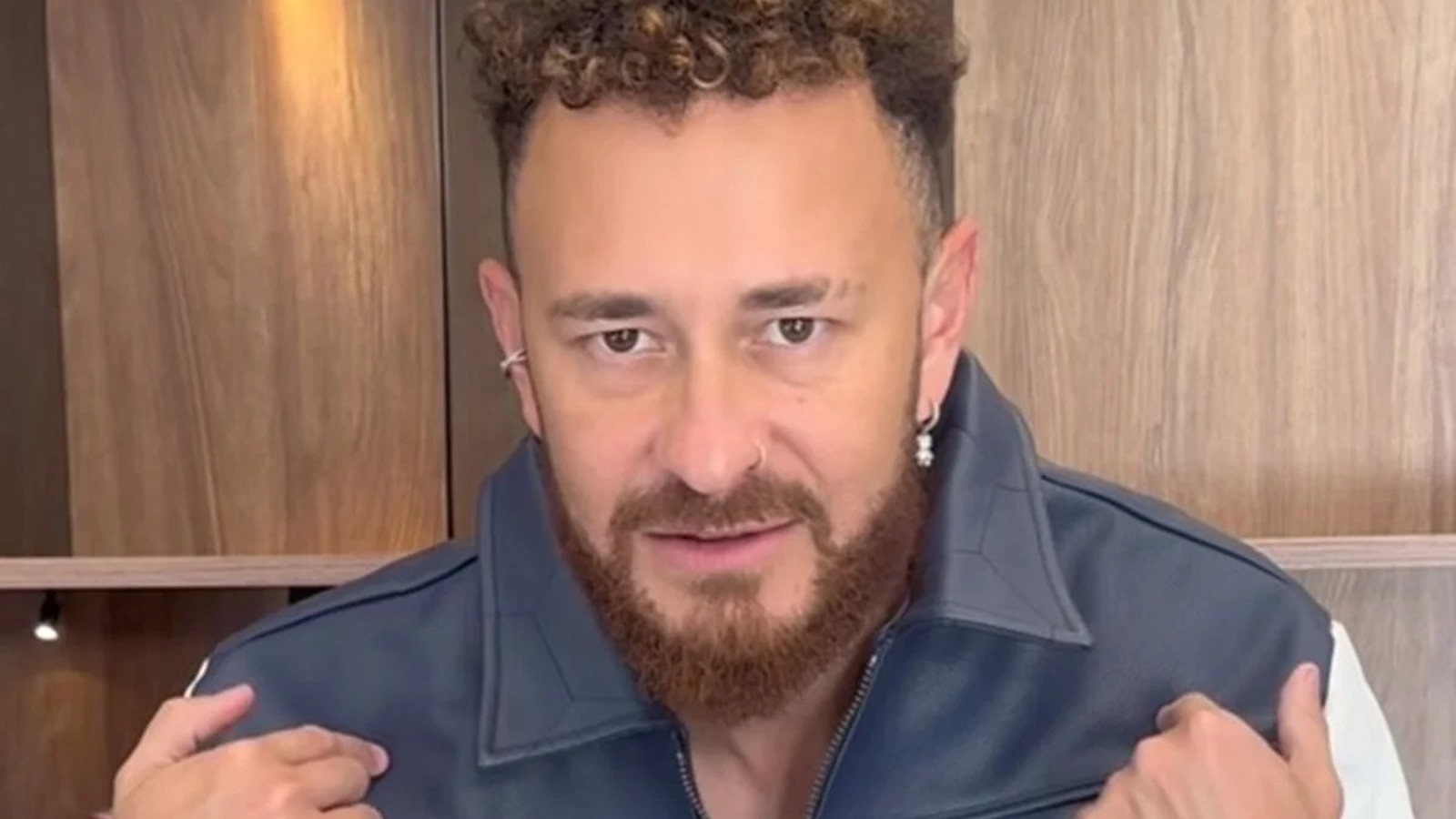 Homem com cabelo encaracolado e barba, vestindo jaqueta azul e branca, olhando para frente