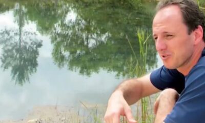 Rogério Ceni de camisa azul à beira da água, com reflexos de árvores e vegetação