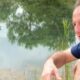 Rogério Ceni de camisa azul à beira da água, com reflexos de árvores e vegetação