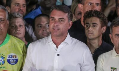 Flávio Bolsonaro em camisa branca, cercado por apoiadores, um com broche político Flávio Bolsonaro