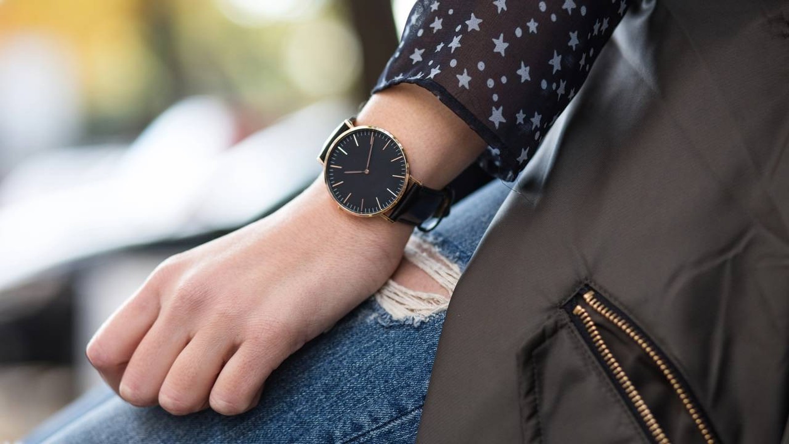 Mão e pulso de pessoa com relógio preto minimalista, camisa de estrelas e jeans rasgado