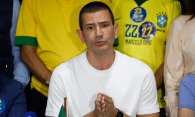 Homem de camiseta branca, mãos juntas. Fundo com camisas amarelas '22 Marcelo Dino' e pin 'Bolsonaro'