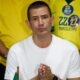 Homem de camiseta branca, mãos juntas. Fundo com camisas amarelas '22 Marcelo Dino' e pin 'Bolsonaro'