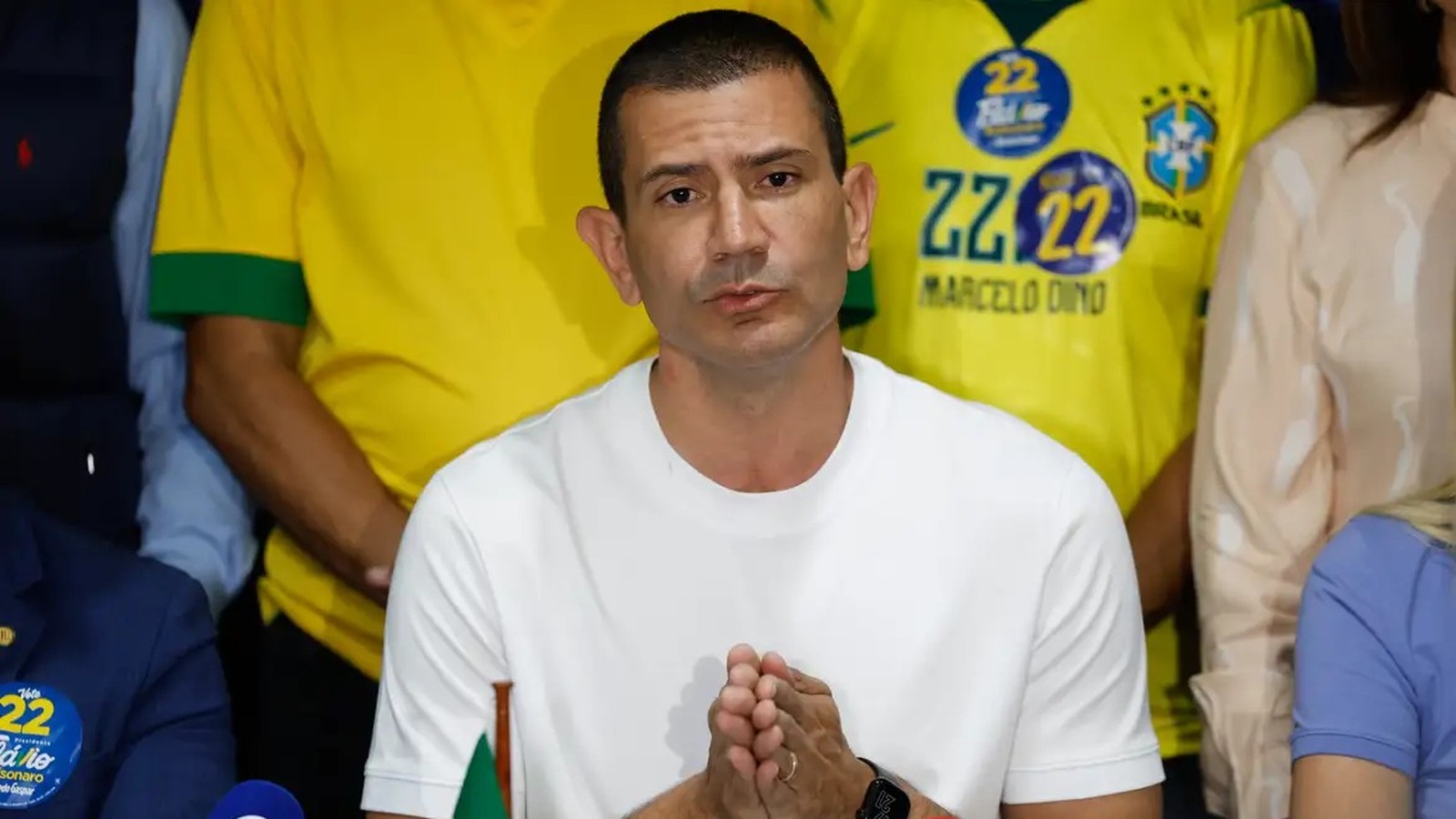 Homem de camiseta branca, mãos juntas. Fundo com camisas amarelas '22 Marcelo Dino' e pin 'Bolsonaro'