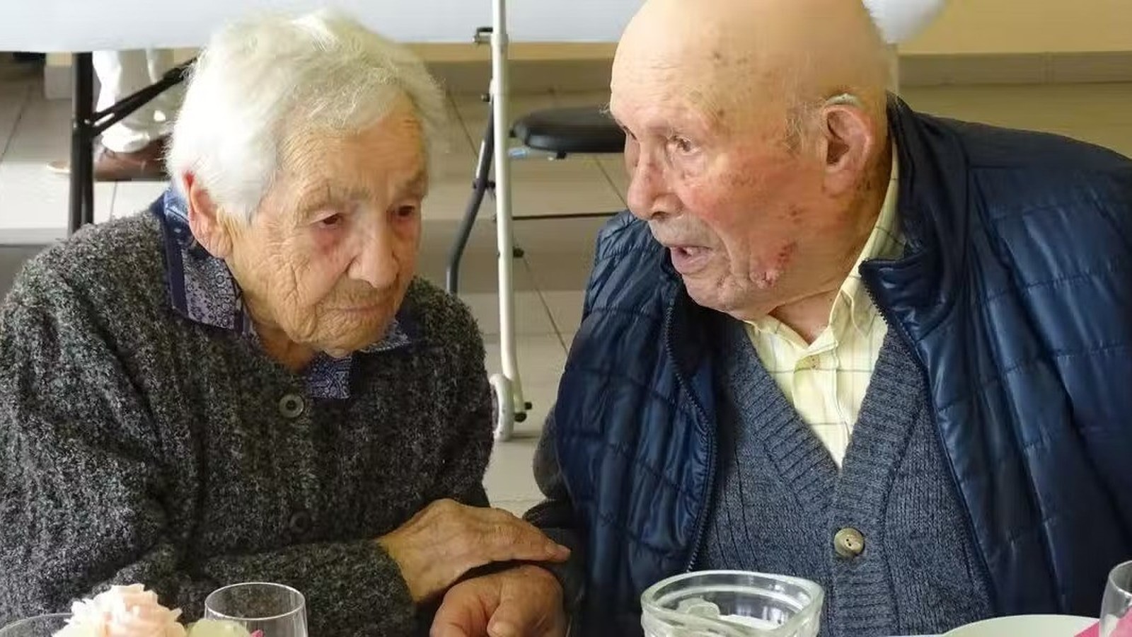 Madeleine e Justin, idosos, sentados à mesa, conversam; ela segura a mão dele.