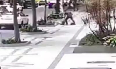 Homem agride pessoa caída no chão na calçada, carros e árvores ao fundo
