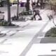 Homem agride pessoa caída no chão na calçada, carros e árvores ao fundo