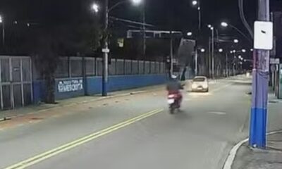 Rua noturna com moto, carro, postes de iluminação e muro azul com letreiro