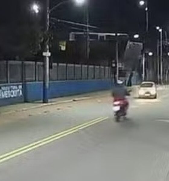 Rua noturna com moto, carro, postes de iluminação e muro azul com letreiro